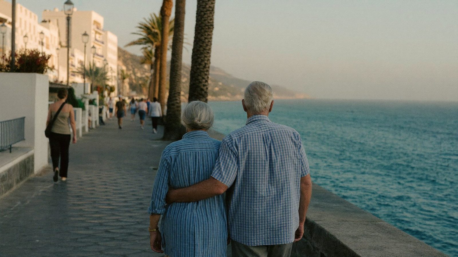 A silver-haired British couple walking arm in arm along a Spanish seafront promenade at golden hour